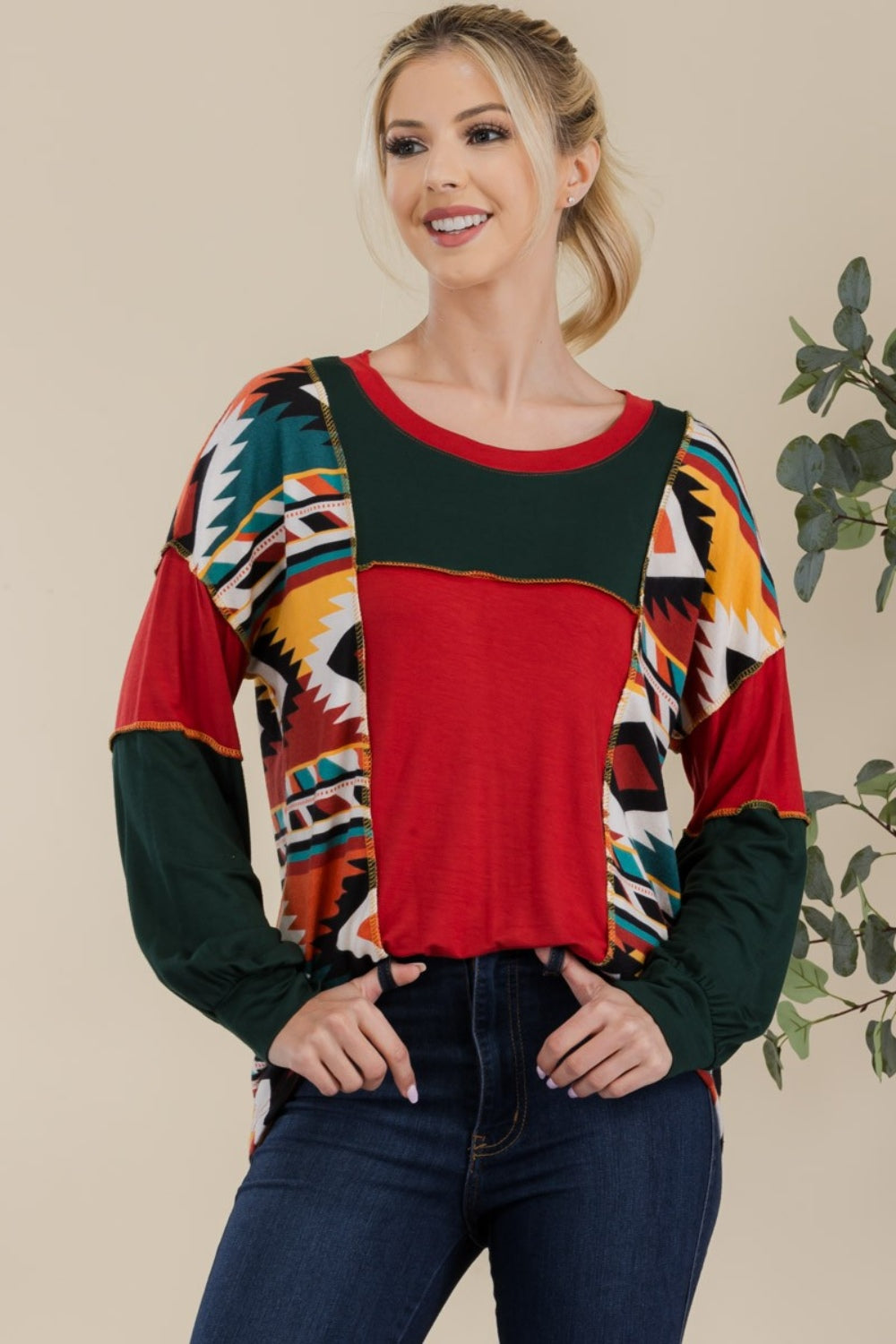 A woman stands against a plain background, wearing the Celeste Full Size Exposed Seam Printed Color Block T-Shirt with vibrant red, green, and yellow geometric sections, paired with dark jeans. She smiles, her hands resting in her jeans' pockets.