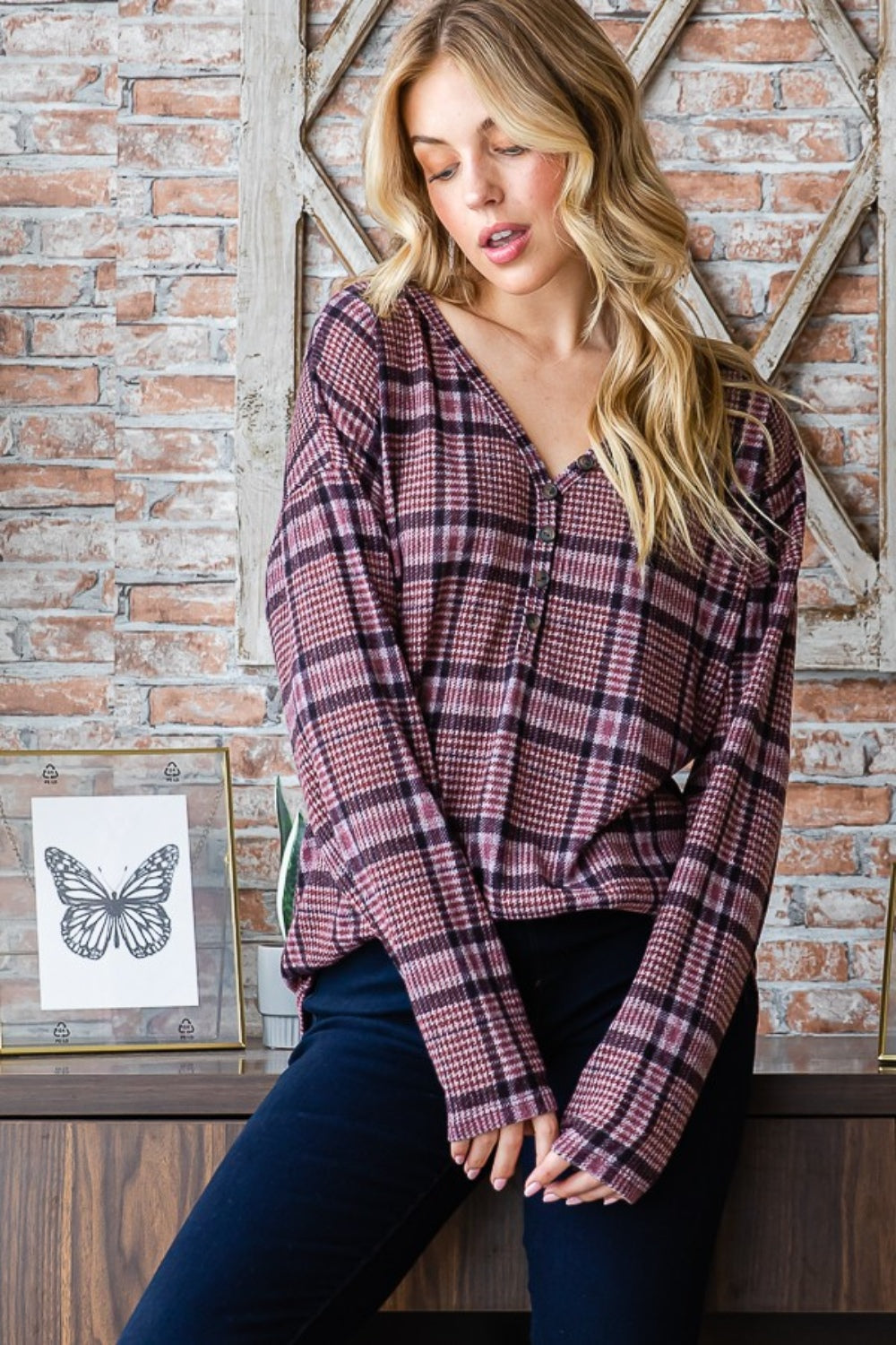 Sporting long blonde hair, a person stands against a brick wall wearing the Heimish Full Size Plaid V-Neck Long Sleeve Top in pink and black. Framed butterfly prints adorn the background, while this adaptable wardrobe item adds a touch of flair to any ensemble.