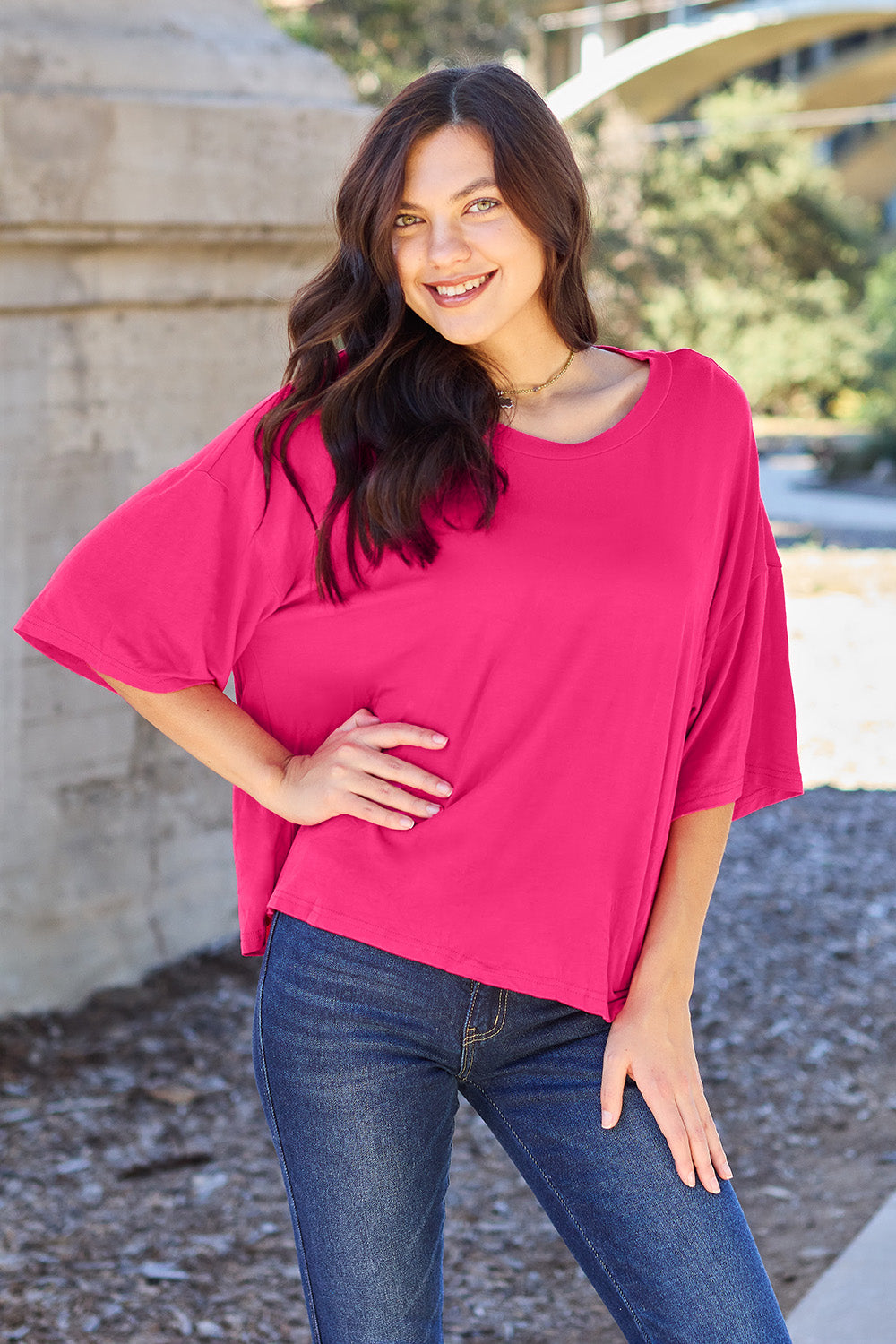 A person with long dark hair and a bright smile stands outdoors, showcasing their basic style in a loose, slightly stretchy *Basic Bae Full Size Round Neck Drop Shoulder T-Shirt* in pink and blue jeans, with one hand on their hip.