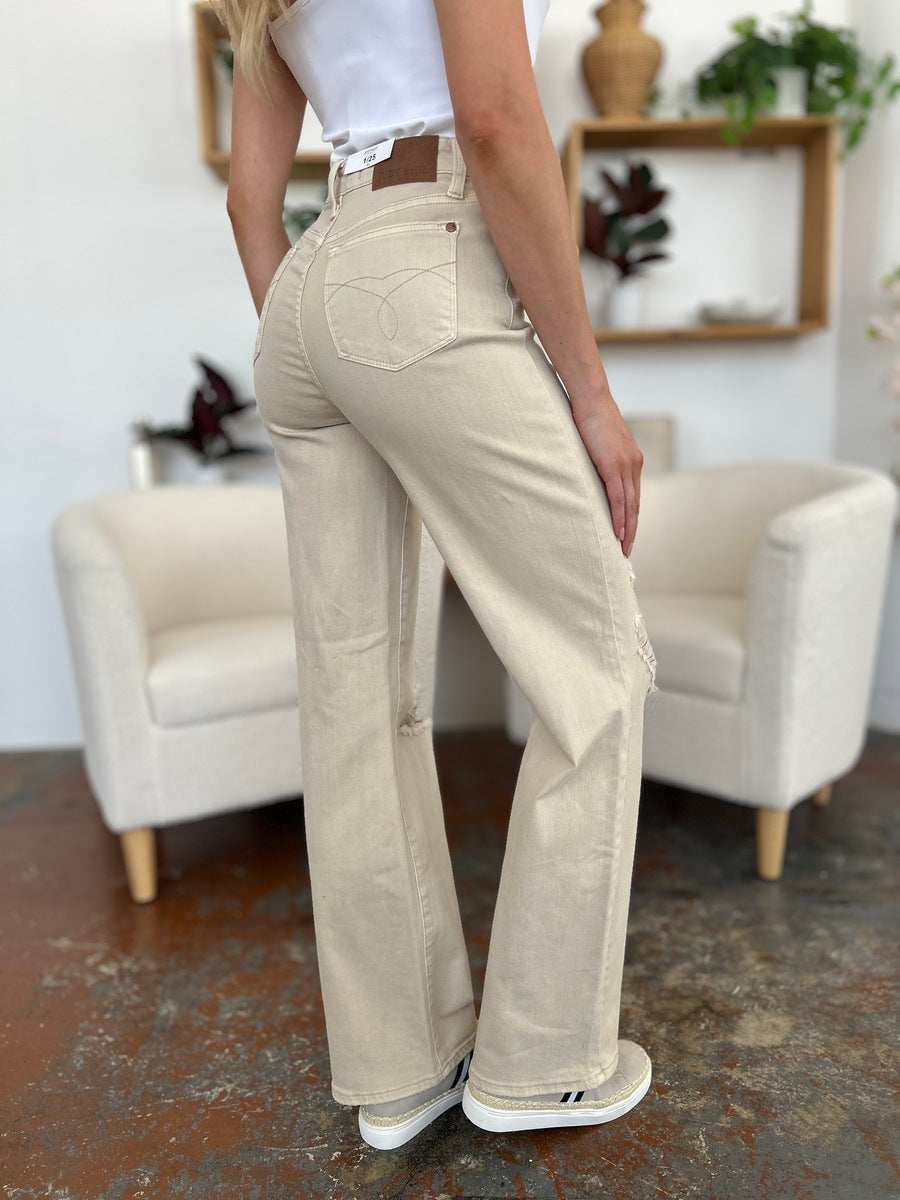 A person stands with hands in pockets, wearing a white shirt, Judy Blue Full Size High Waist Distressed Wide Leg Jeans, and black sandals. The background features a white chair, shelves, and a potted flower.