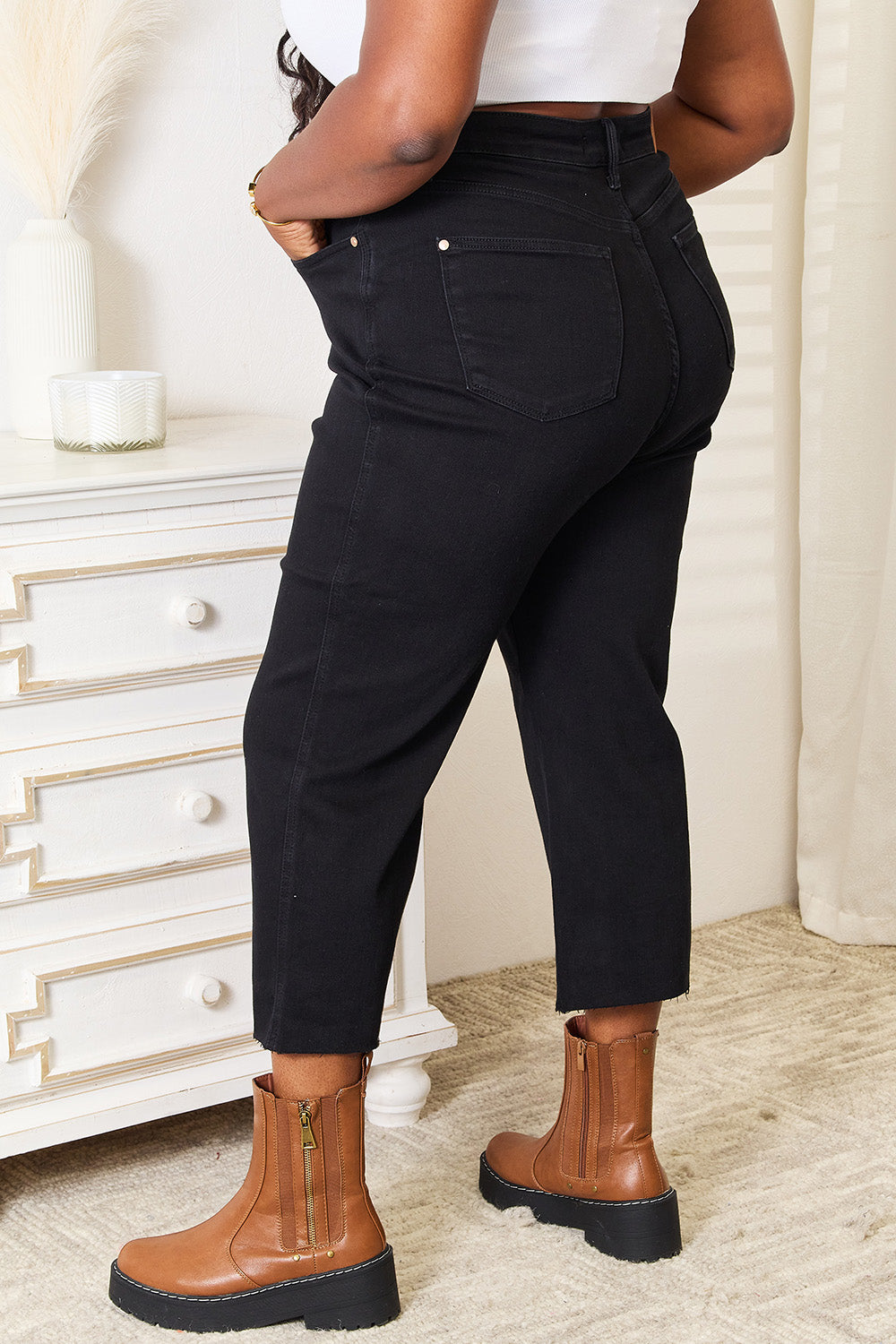 A person wearing Judy Blue Full Size High Waist Wide Raw Hem Cropped Jeans in black and a white lace top stands next to a white dresser adorned with a vase and candle.