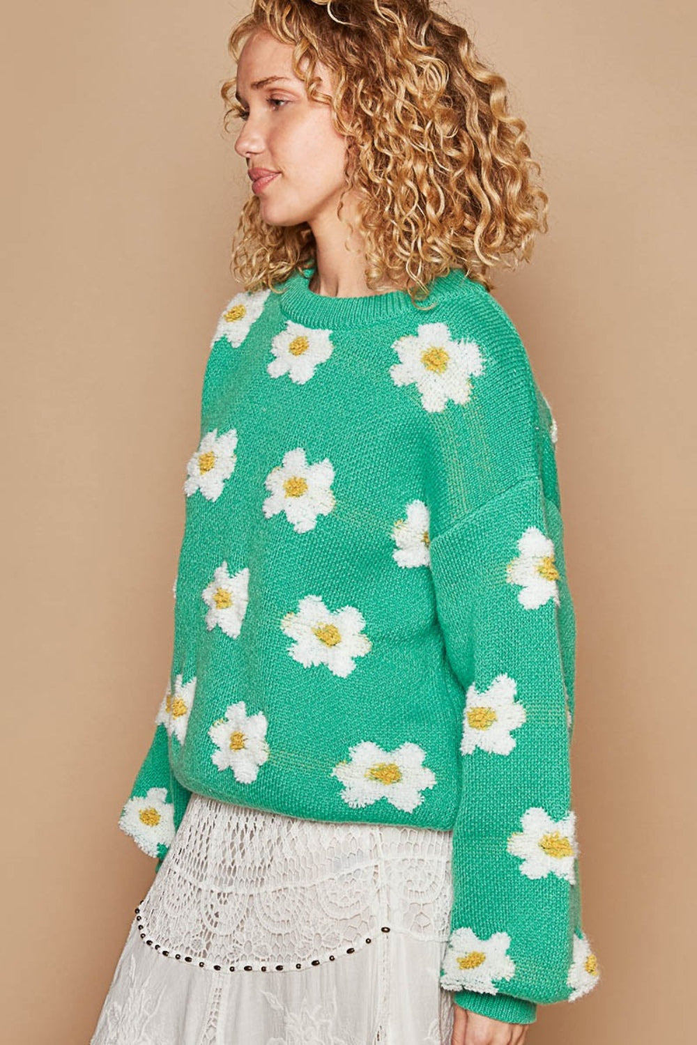 A person with curly hair wearing the POL Daisy Pattern Drop Shoulder Sweater, which is an oversized green sweater with a daisy pattern, and a lace skirt stands against a beige background, smiling.