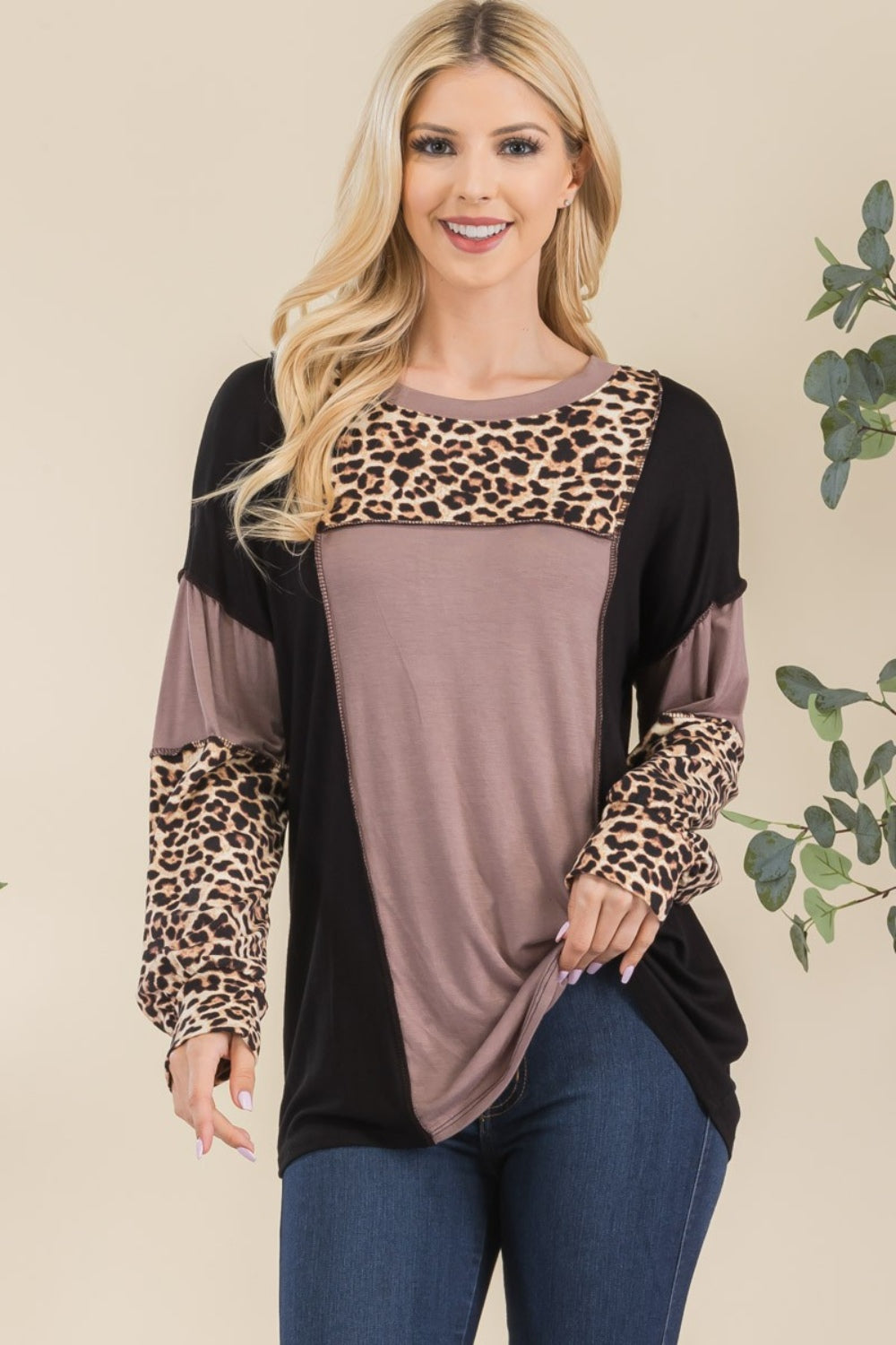 A person wearing the Celeste Full Size Leopard Color Block Exposed Seam T-Shirt and dark jeans stands against a neutral background with greenery. The long-sleeved shirt, with its stylish exposed seams, features cozy leopard print accents, adding subtle flair to this chic ensemble.