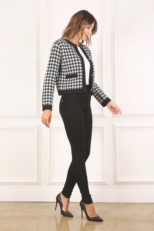 A woman dressed in the Black Check Knitted Jacket, featuring snap buttons and functional pockets, complemented by black pants, stands against a white background.