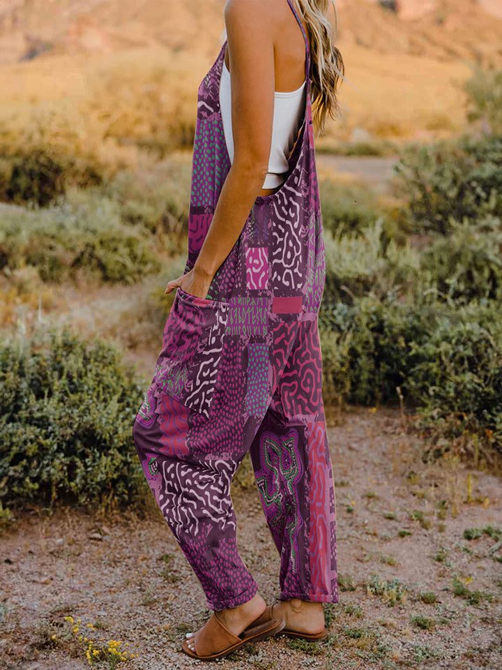 A person standing outdoors in front of rocky terrain, wearing a Full Size Printed V-Neck Sleeveless Jumpsuit and a hat.