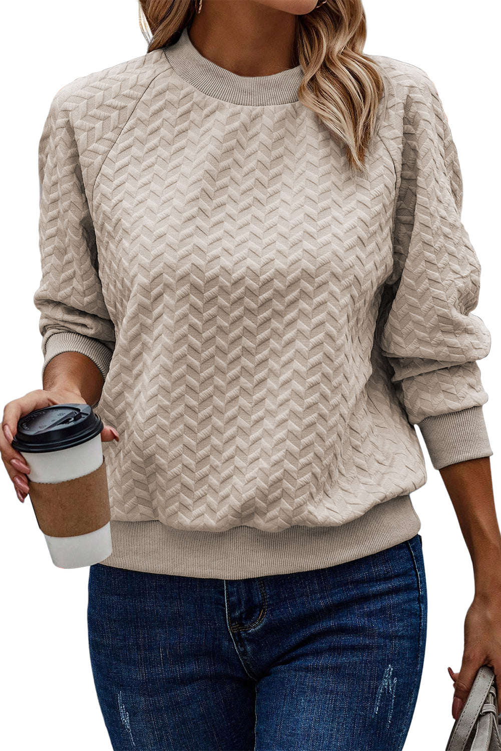 A woman in a Beige Solid Textured Raglan Sleeve Pullover Sweatshirt and blue jeans is looking to the side. She sports a brown hat and earrings, exuding a casual and comfy vibe. Standing next to dried pampas grass against a light background, she seamlessly blends with her textured sweatshirt.