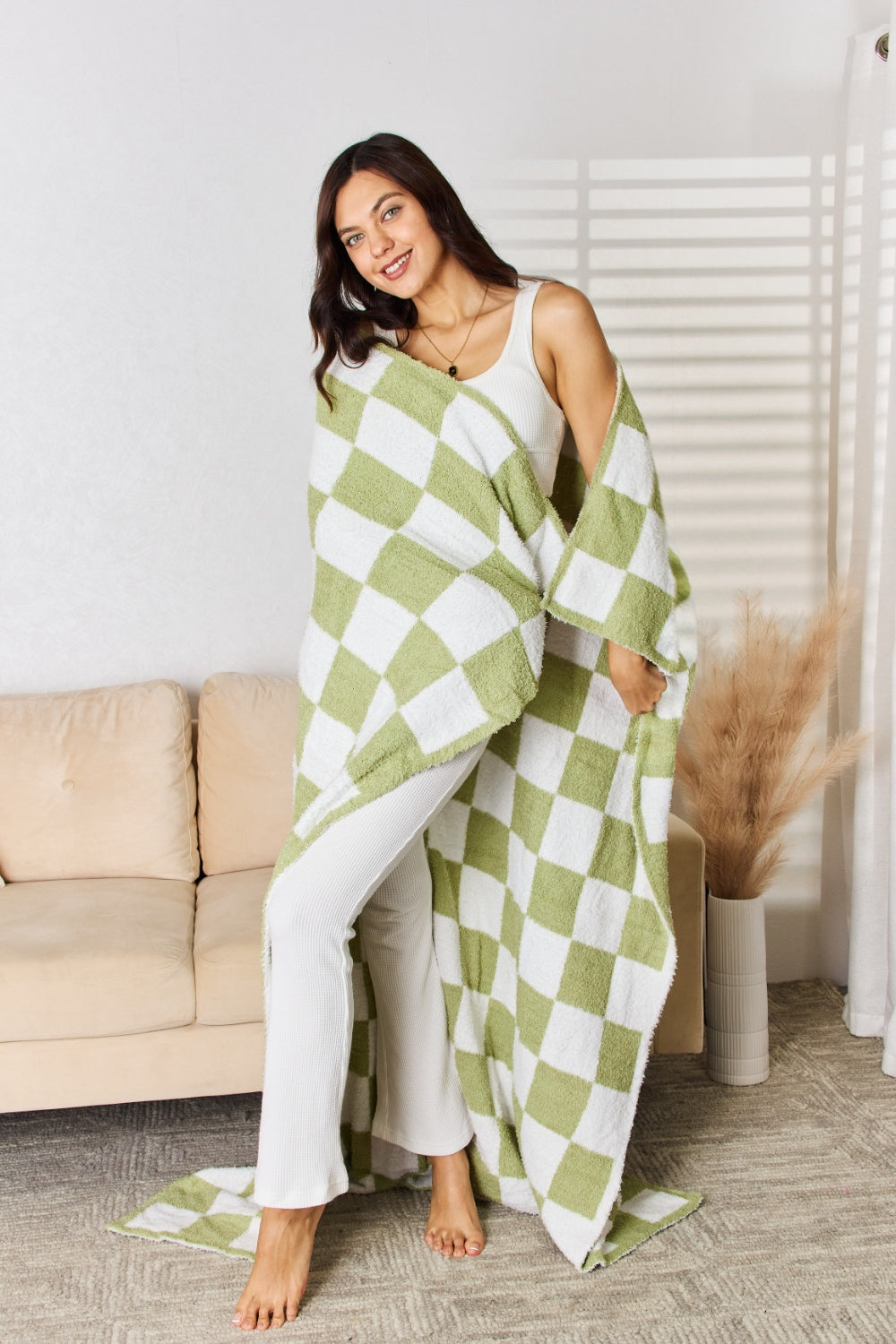 A person stands holding a large Cuddley Checkered Decorative Throw Blanket, smiling, in a room with a sofa and a potted plant. The blanket features a black and white checkerboard pattern, is made from 100% polyester, and imported for quality.