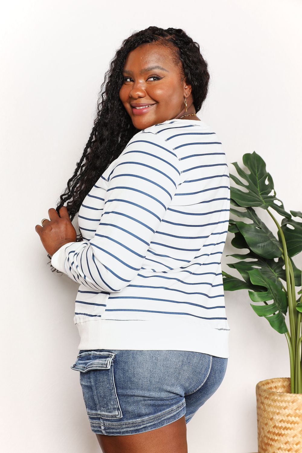 A person smiling while wearing the Double Take Striped Long Sleeve Round Neck Top, paired with denim shorts, stands in front of a white background with a plant.