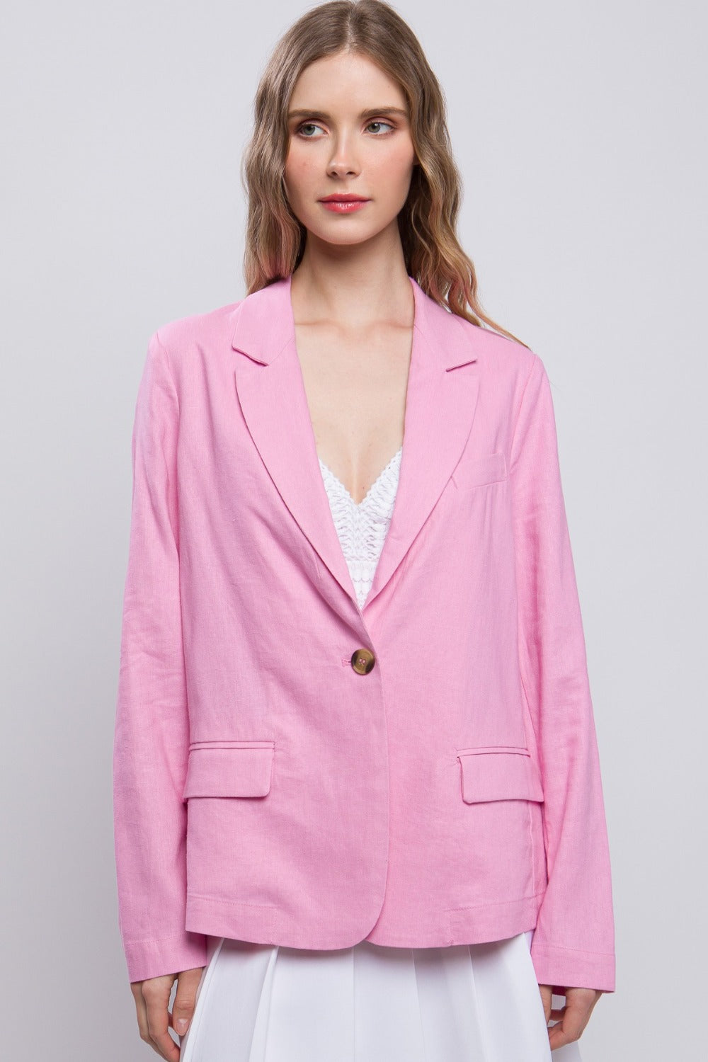 A person with long hair wears a pink Love Tree One-Button Long Sleeve Linen Blazer over a white top, posing with one hand touching their hair.