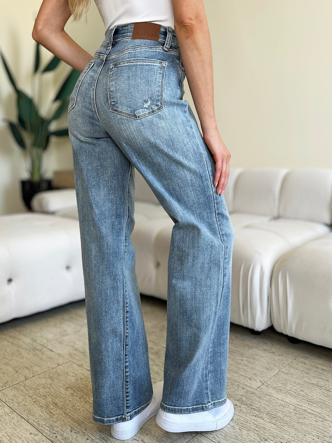 A person wearing Judy Blue Full Size High Waist Straight Jeans and white shoes is standing indoors. There is a white couch and a large plant in the background.