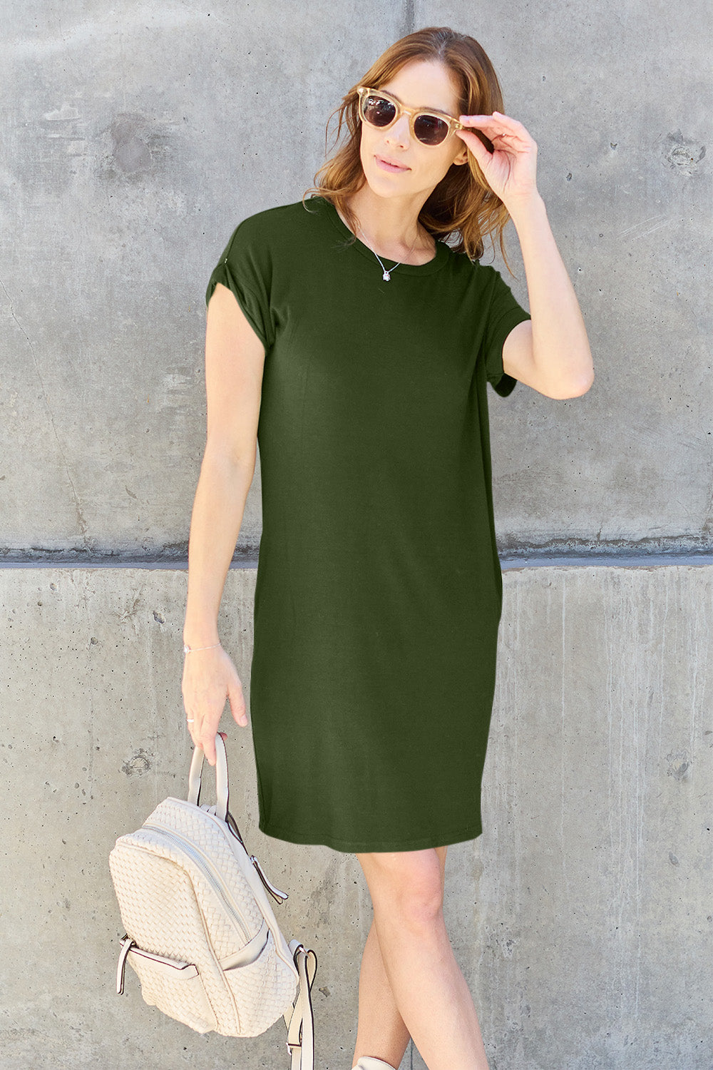 A woman wearing a dark olive-green Basic Bae Full Size Round Neck Short Sleeve Dress with Pockets and sunglasses stands against a concrete wall, holding a light-colored backpack.