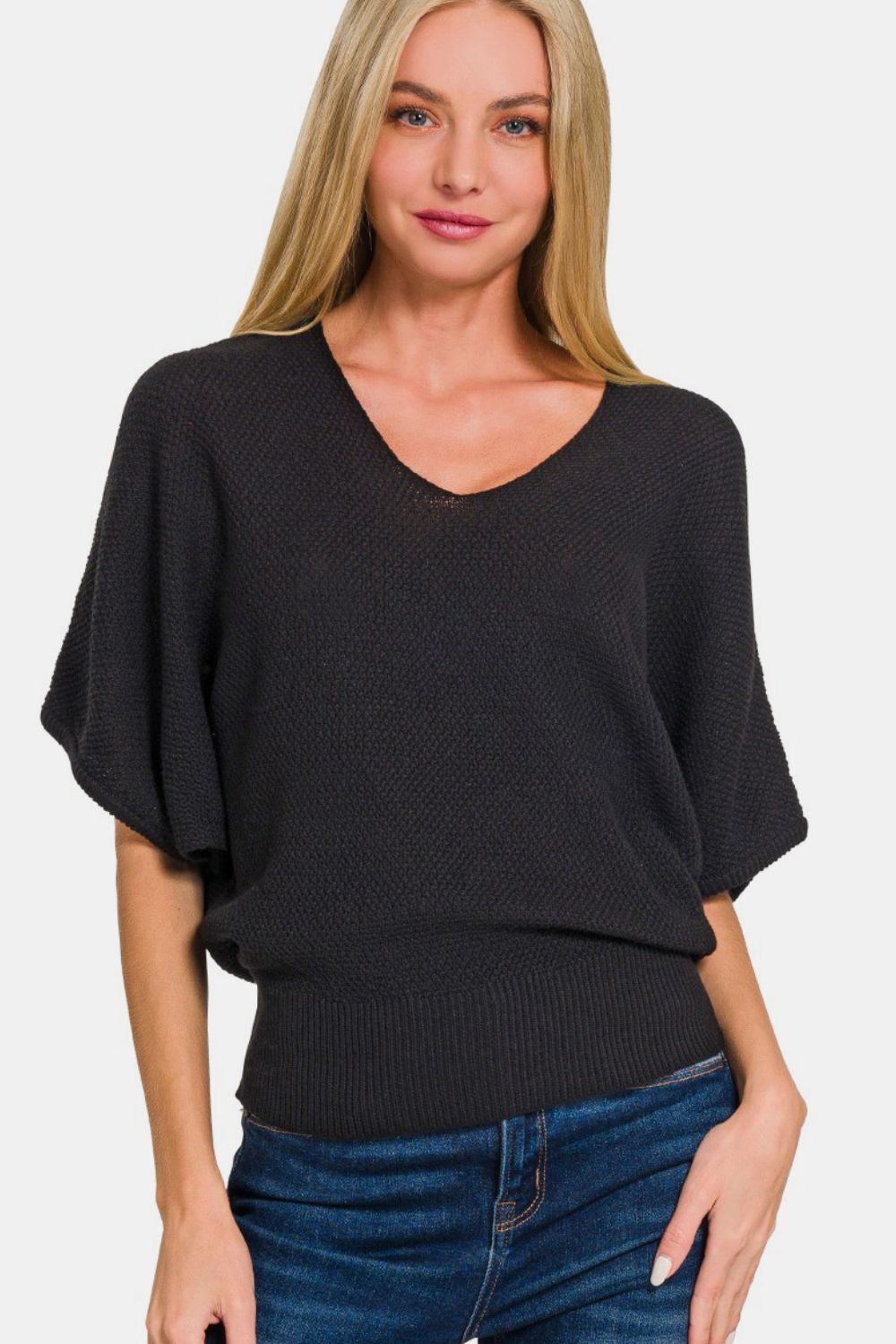 A person with long, blonde hair is wearing a Zenana V-Neck Short Sleeve Dolman Sweater in black and blue jeans, standing against a plain white background.