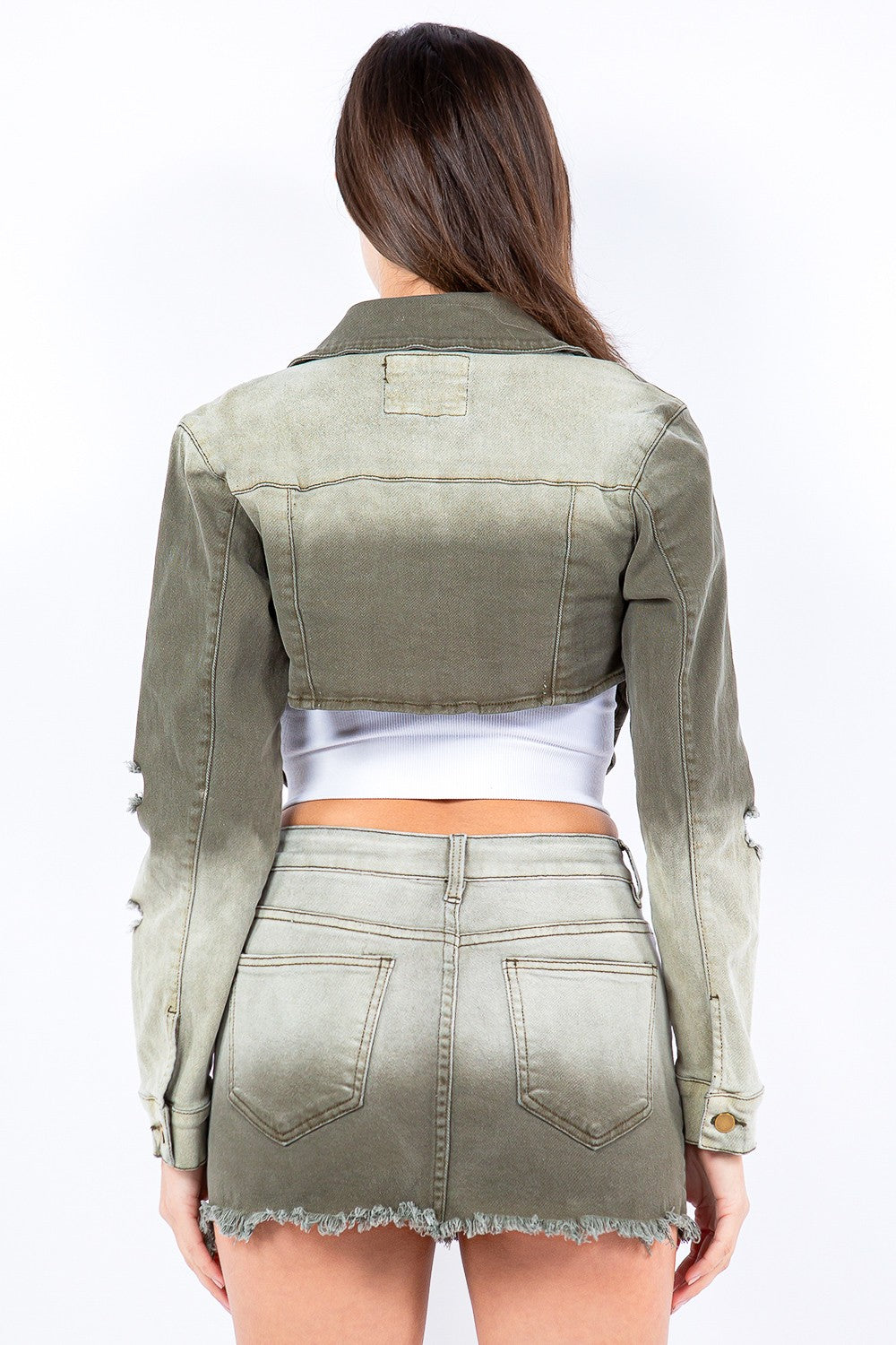 A person stands wearing an American Bazi Distressed Ombre Washed Cropped Denim Jacket in light olive green, paired with a matching skirt featuring a frayed hem and a white cropped tank top.