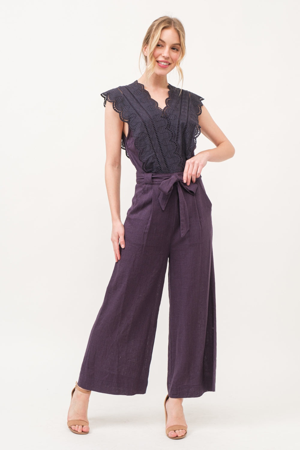 A person in an elegant and fashionable And The Why Laced Surplice Tie Waist Jumpsuit with wide-leg pants stands against a plain background, smiling with hands on hips.