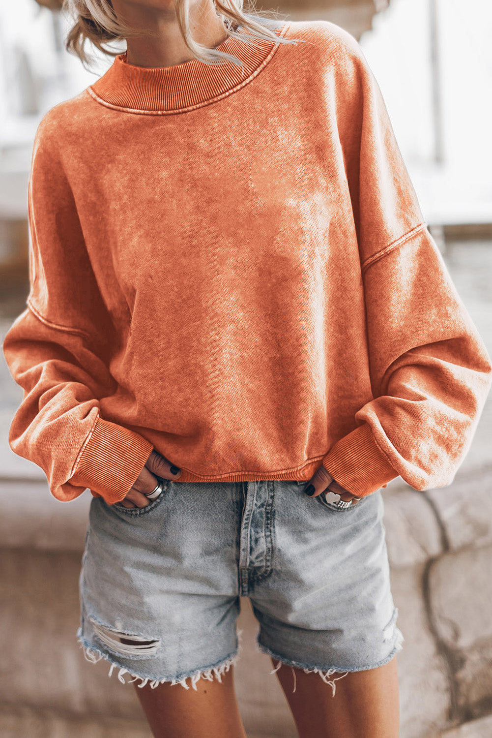 A person with long blonde hair wearing an Orange Drop Shoulder Crew Neck Pullover Sweatshirt and blue jeans stands with their back to the camera in a sunflower field.