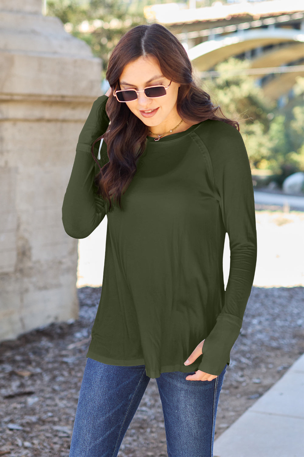 A woman wearing sunglasses and a slightly stretchy Basic Bae Full Size Round Neck Long Sleeve T-Shirt in white stands on a sidewalk with an arched structure in the background, smiling slightly.