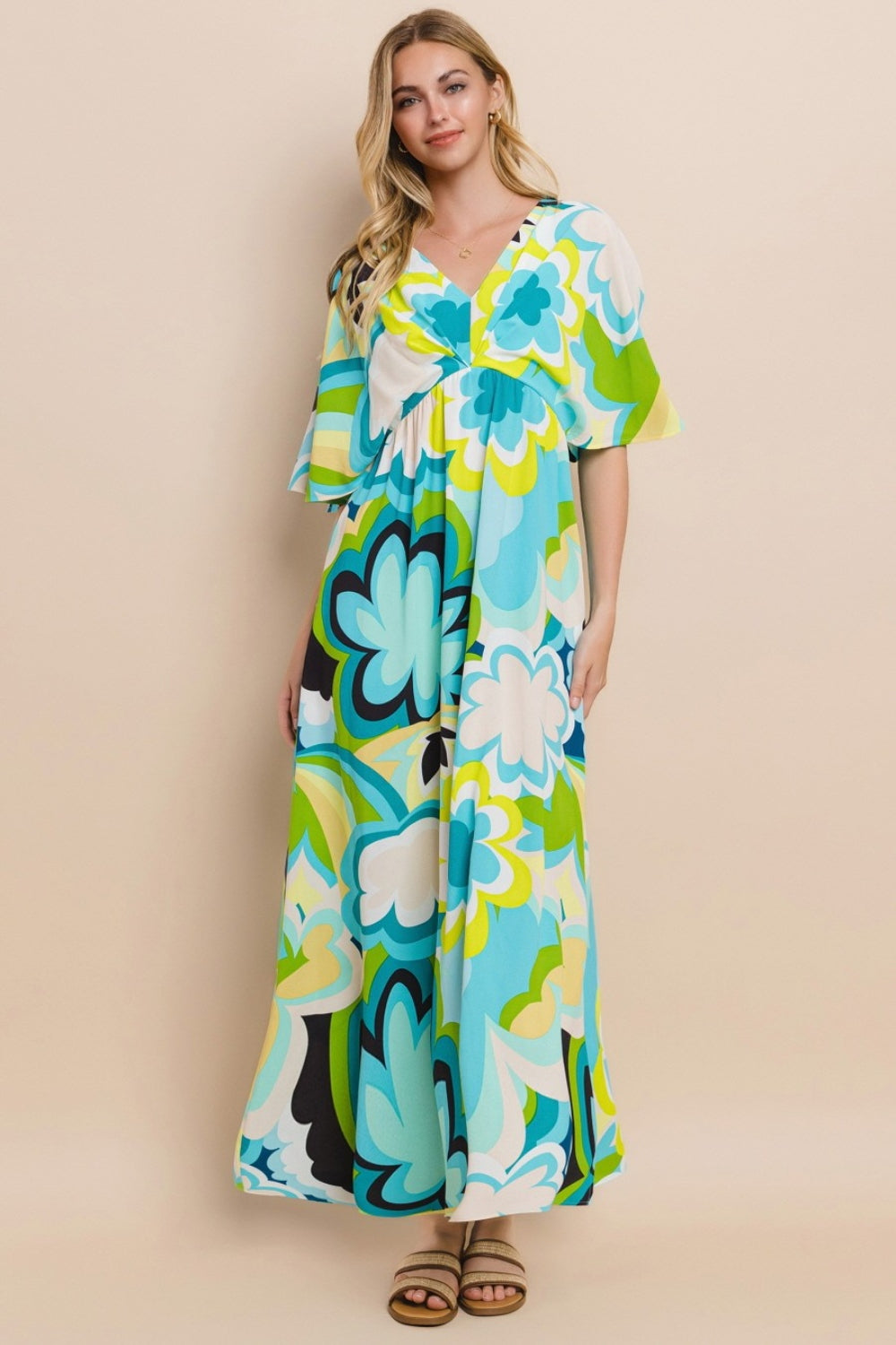 Against a simple backdrop, a woman models the ODDI Floral Printed Slit Maxi Dress, exuding a sense of versatile elegance. This maxi dress embodies summer chic with its vibrant floral design featuring shades of blue, green, and yellow.