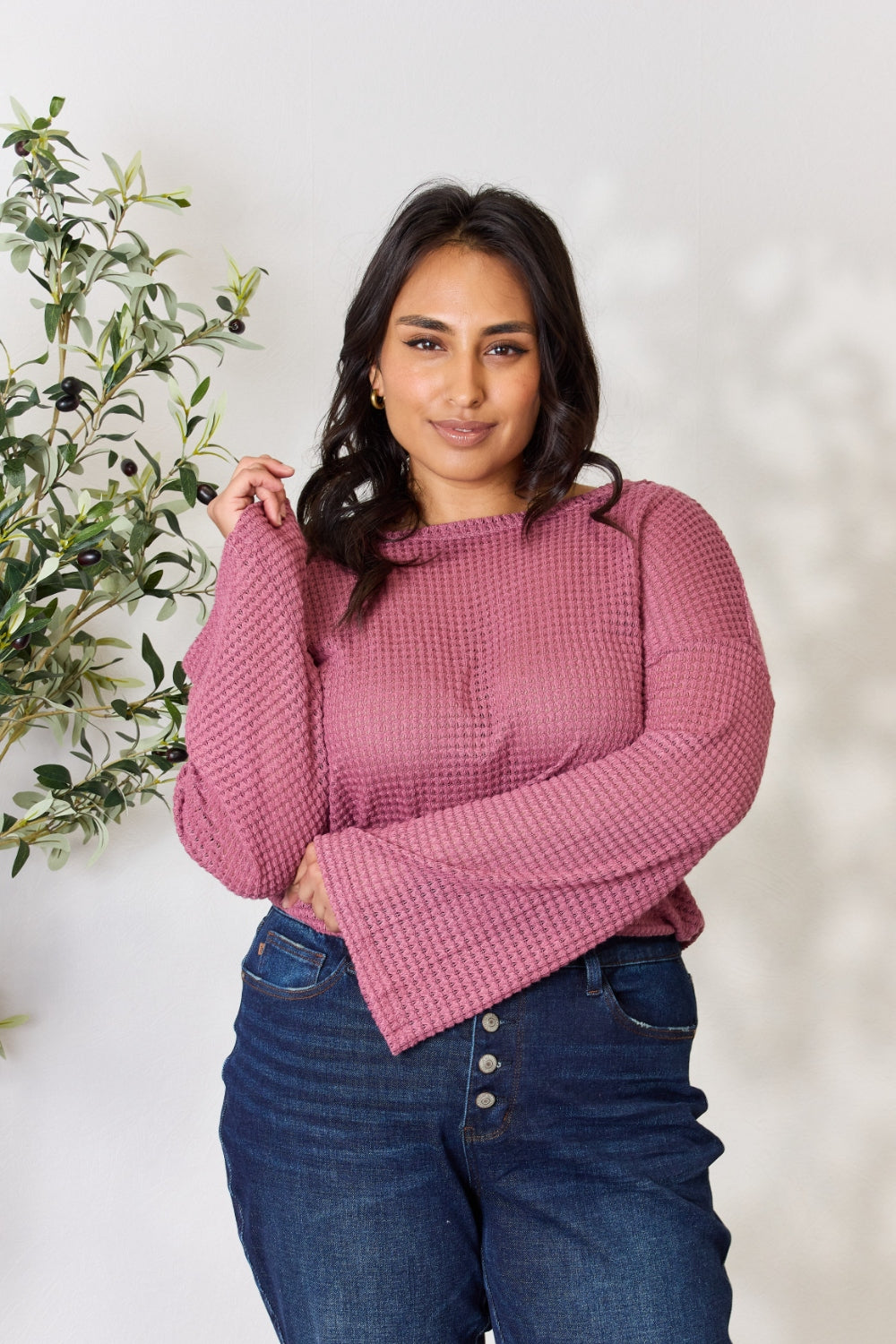 A person wearing a Culture Code Full Size Waffle-Knit Round Neck Long Sleeve Blouse in pink and jeans stands in front of a light background, complemented by a leafy plant on the left.