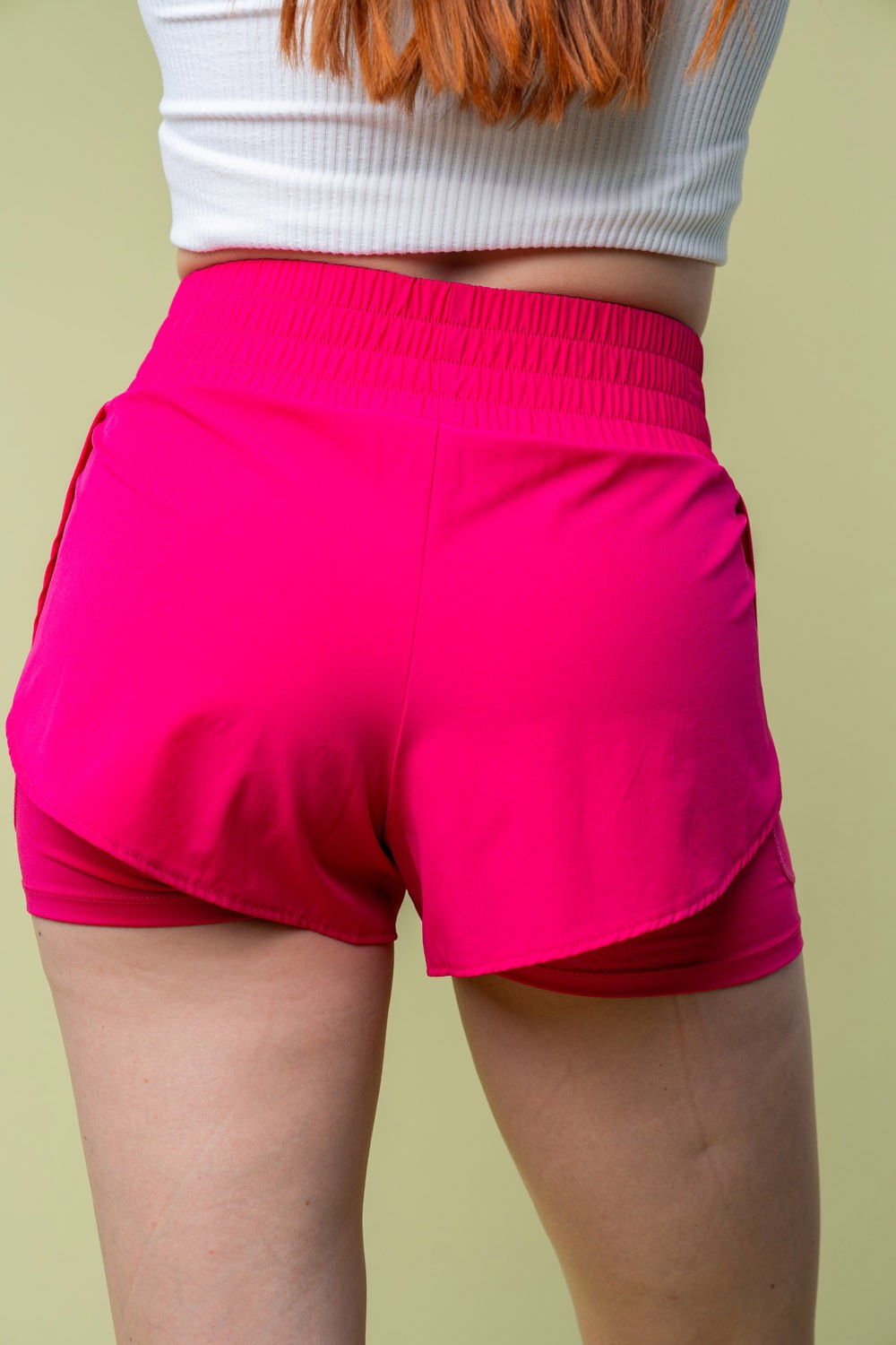 A person wearing White Birch Full Size High Waisted Knit Shorts in bright pink and a white cropped top stands against a light green background.