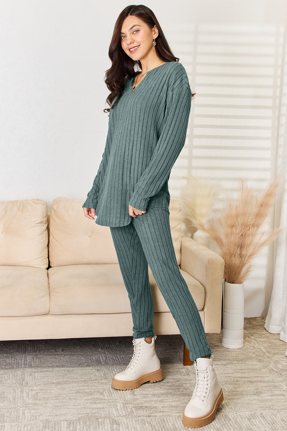 An individual is seen indoors sporting the Basic Bae Full Size Notched Long Sleeve Top and Pants Set, a matching red two-piece outfit that includes a long-sleeve top and pants, complemented by tan high-heeled sandals. A decorative ladder and pampas grass in a vase are visible in the background. This slightly stretchy ensemble ensures both comfort and style and is machine washable with cold water.