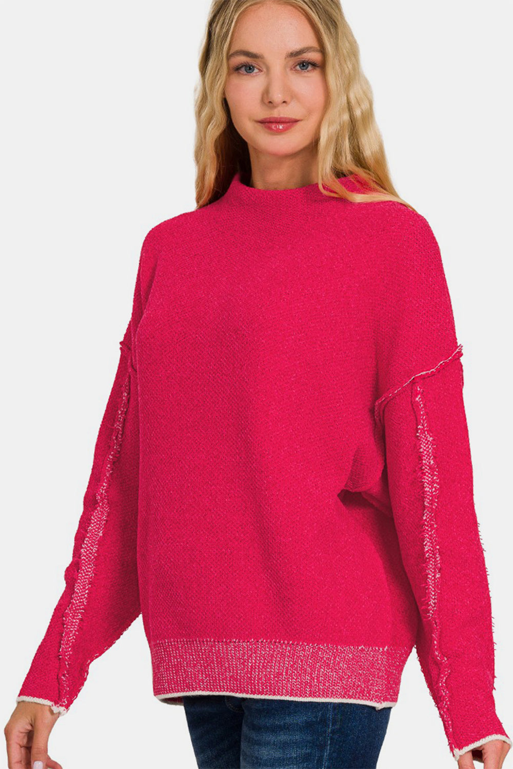 A woman in a vibrant pink Zenana Exposed Seam Mock Neck Long Sleeve Sweater and jeans stands elegantly against a minimalist white background.