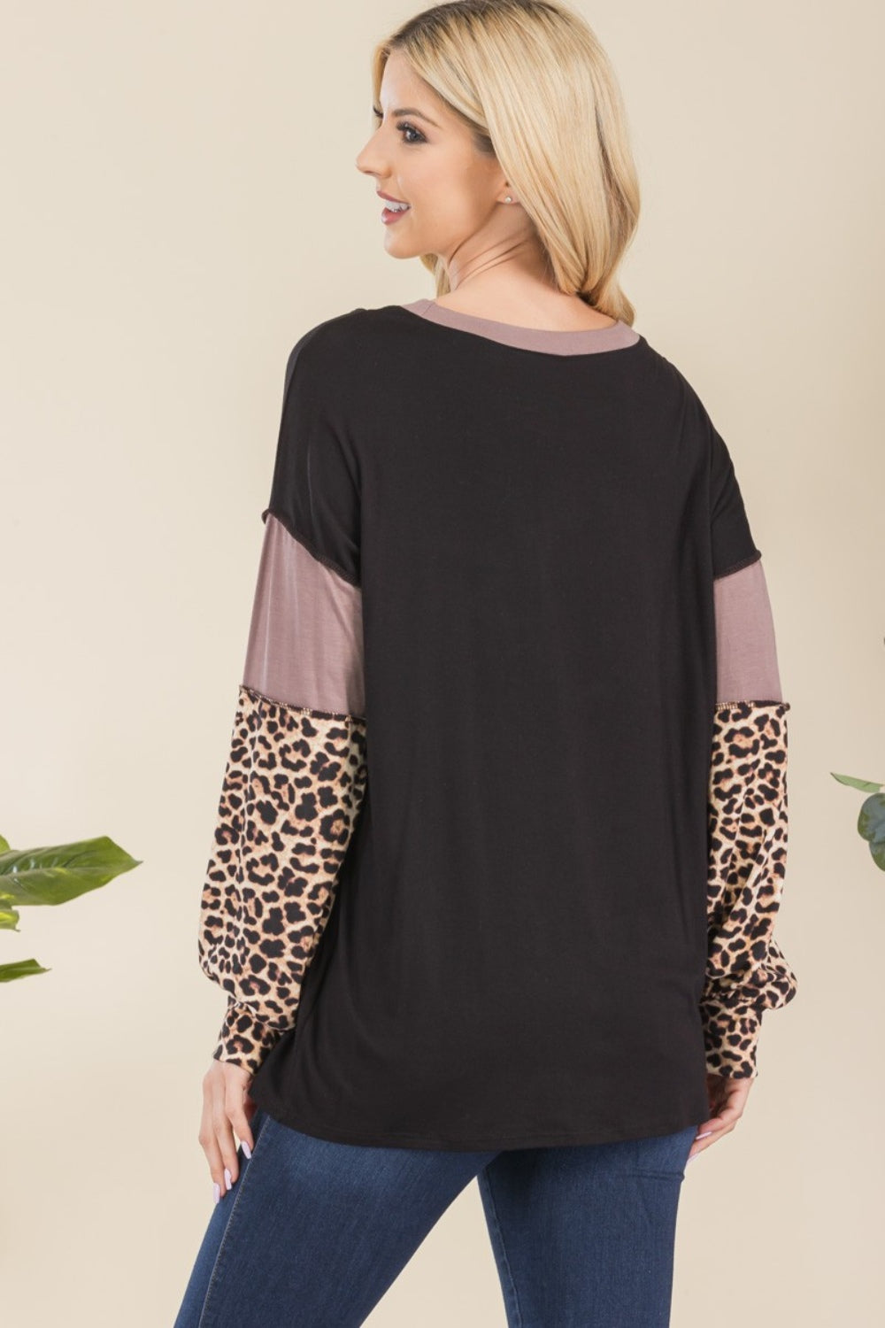 A person wearing the Celeste Full Size Leopard Color Block Exposed Seam T-Shirt and dark jeans stands against a neutral background with greenery. The long-sleeved shirt, with its stylish exposed seams, features cozy leopard print accents, adding subtle flair to this chic ensemble.