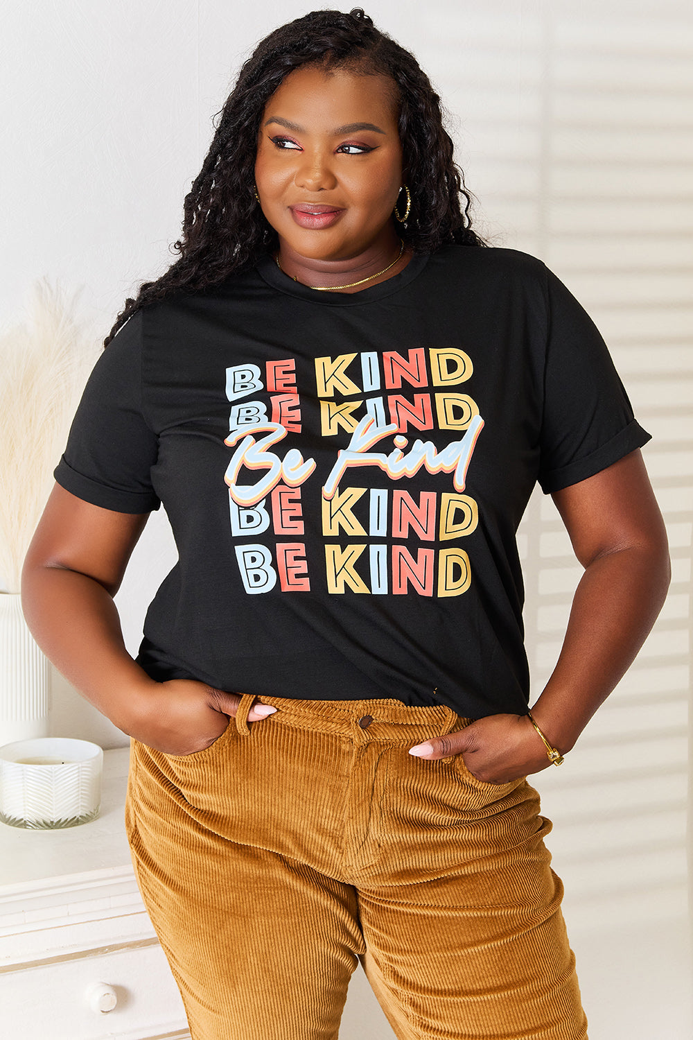 A woman stands with her hands in the pockets of her brown corduroy pants, wearing a casual Simply Love BE KIND Graphic Round Neck T-Shirt that reads "BE KIND" in large colorful letters, showcasing positive message clothing.