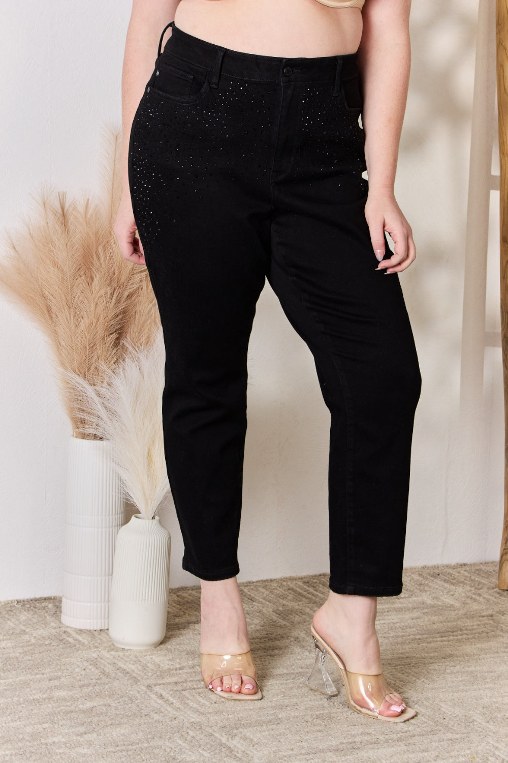 A person wearing slightly stretchy, black Judy Blue Full Size Rhinestone Embellished Slim Jeans and black shoes stands on a textured floor next to a white vase with brown and white ornamental grasses and a wooden ladder.