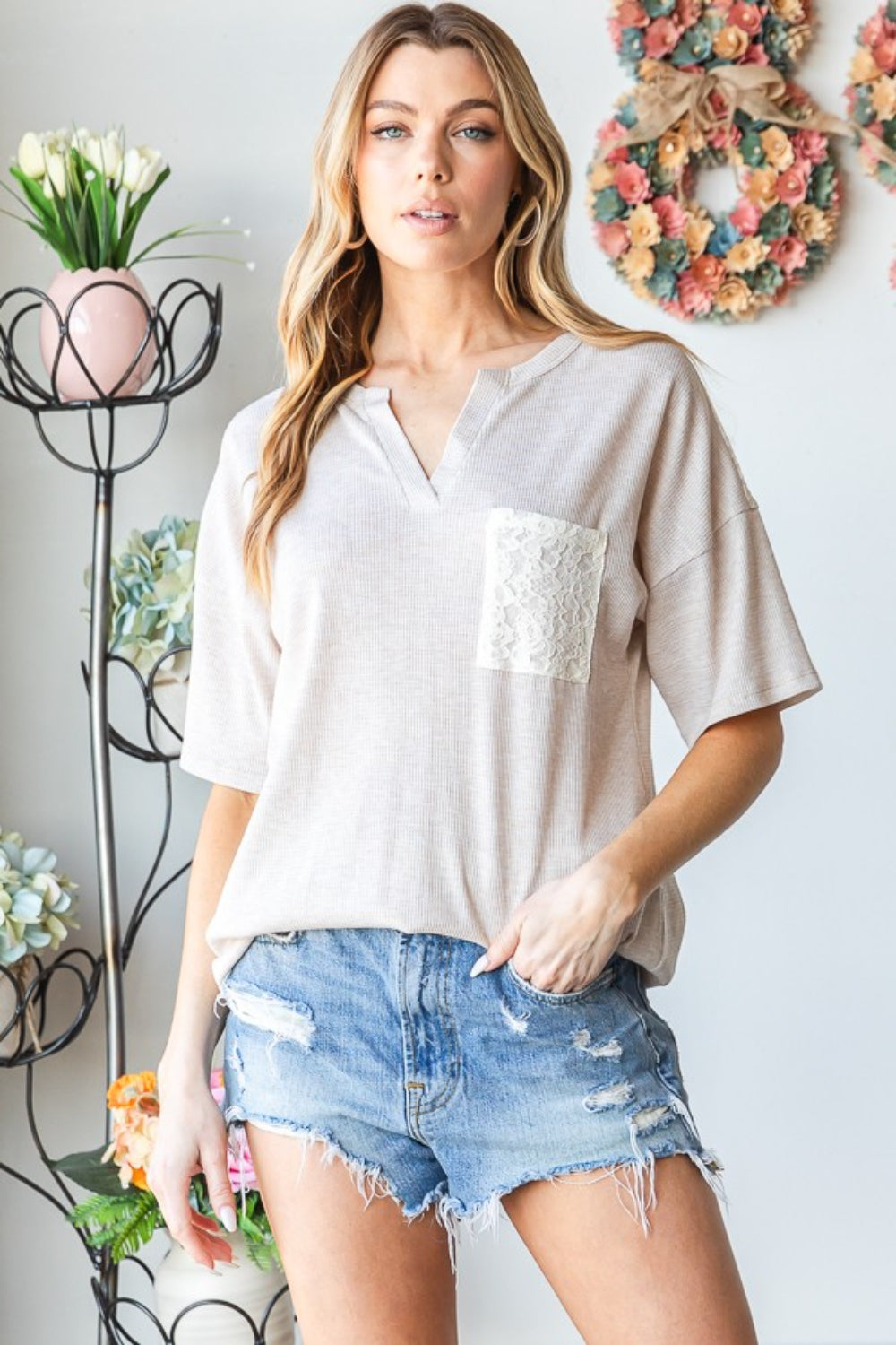 A woman wearing the Heimish Full Size Lace Front Pocket Drop Shoulder Top stands indoors near a decorative metal stand adorned with flowers.