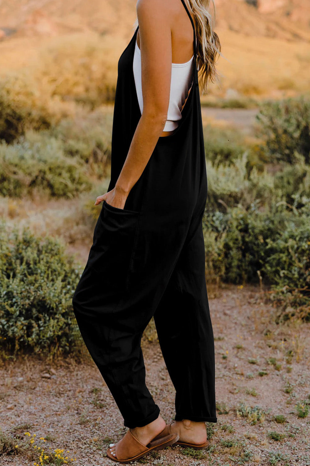 Person wearing a Double Take Full Size V-Neck Sleeveless Jumpsuit with Pockets and sandals, walking outdoors on a dirt trail with bushes in the background.