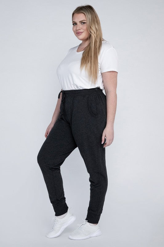 A person stands against a white background, wearing a white t-shirt, grey Plus-Size Jogger Pants with an adjustable waistband, and white sneakers. They have long blonde hair and are posing with one hand on their hip.