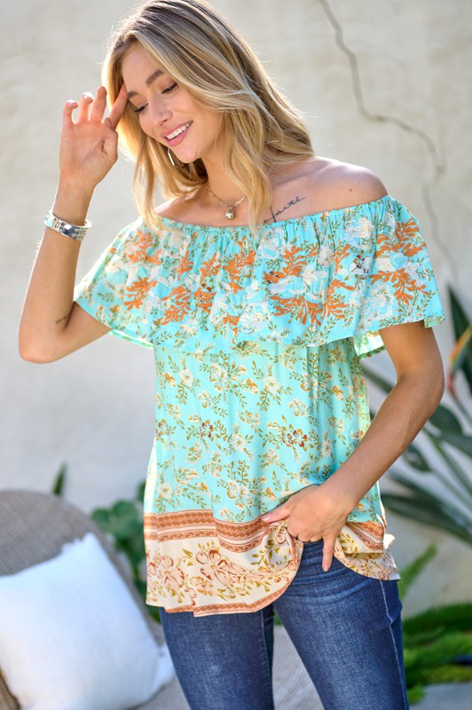 A woman with blonde hair is smiling while standing outdoors, wearing the Printed Off Shoulder Smocked Top paired with jeans.