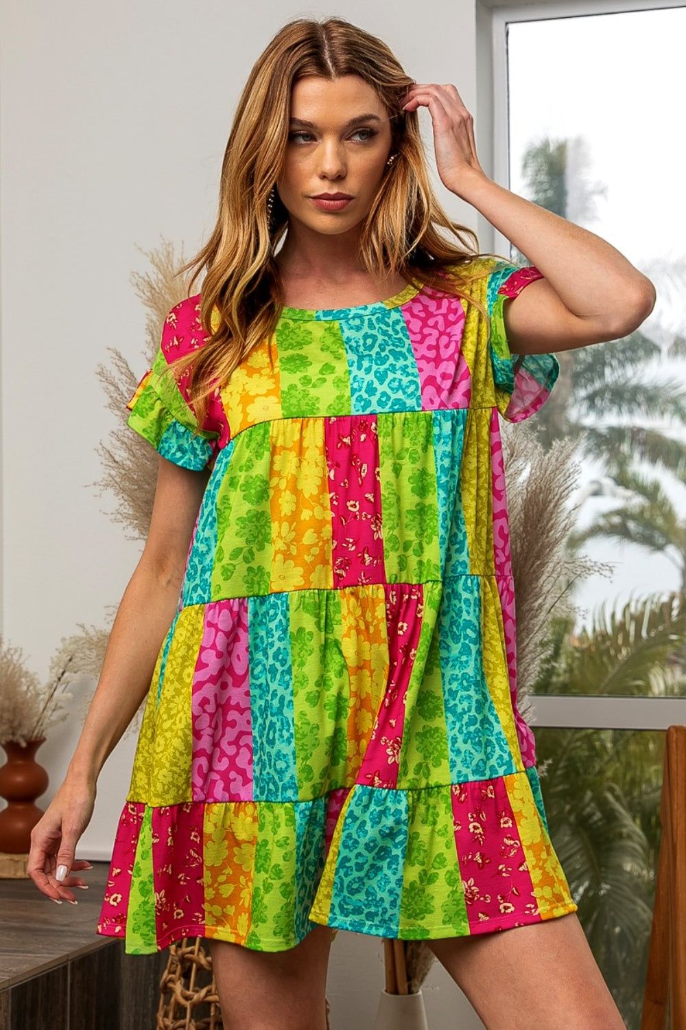 A woman in a BiBi Leopard Short Sleeve Tiered Dress poses with her arms raised behind her head. She stands indoors near a window, framed by pampas grass decor, exuding effortless style and grace.