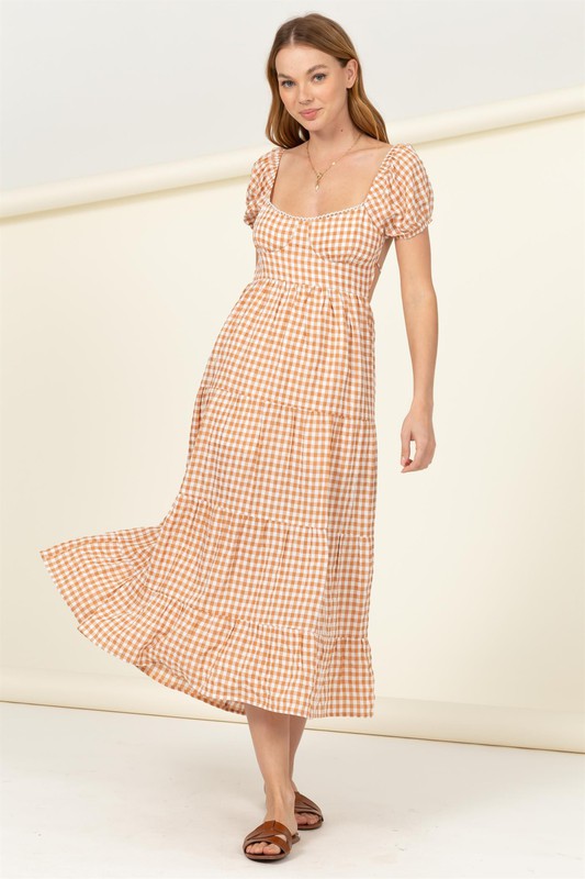 Wearing the "Somewhere to Go Tie-Back Gingham Print Maxi Dress," which features a black and white gingham print with puffed sleeves, a tiered skirt, and elegant tie-back detail, a person poses stylishly paired with black sandals.