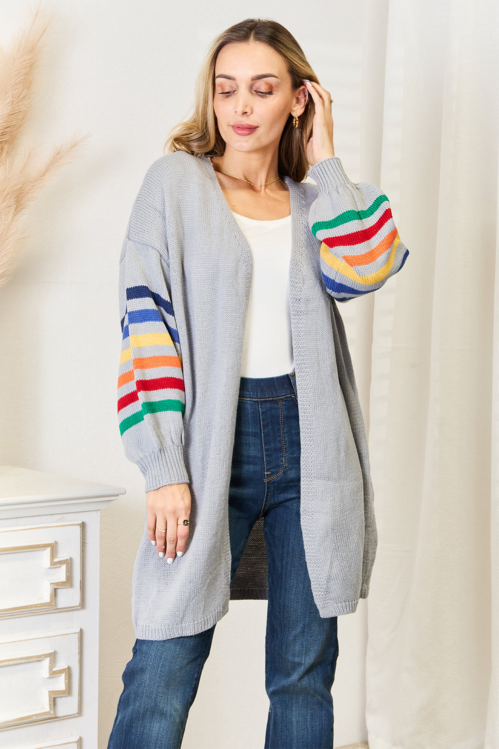 A person wearing the Angel Wings Multicolored Stripe Open Front Longline Cardigan over a white top and blue jeans stands indoors near a white cabinet, showcasing a fashionable cozy look.