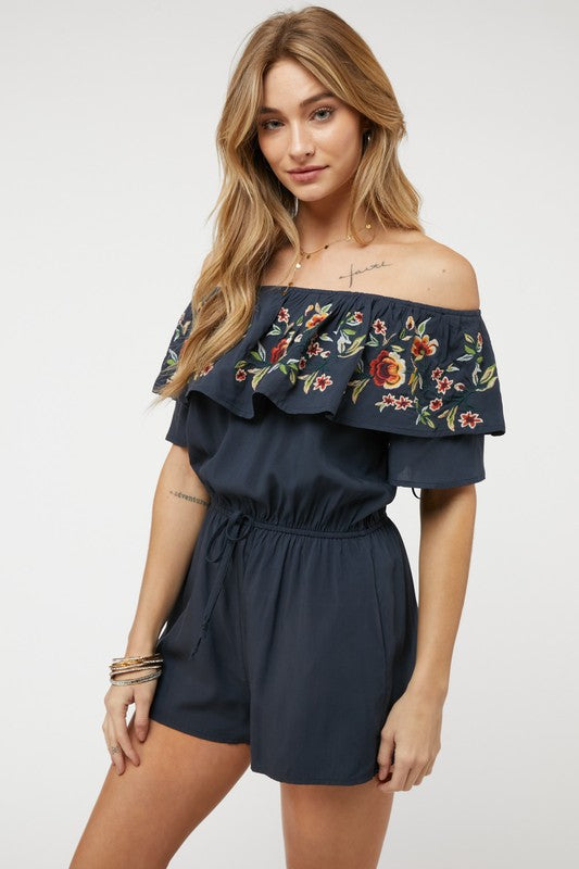 Wearing the stylish Floral Embroidered Off Shoulder Romper, a woman poses gracefully, showcasing its ruffle detail and making it an ideal addition to any summer wardrobe.