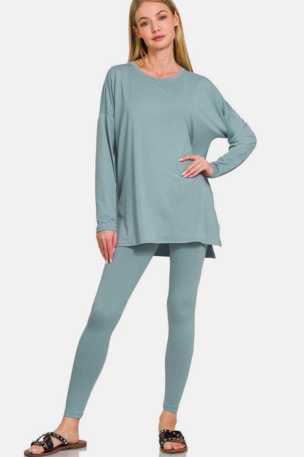 A person wearing the Zenana Full Size Brushed Microfiber Top and Leggings Lounge Set, featuring a loose-fitting teal long-sleeve top and matching stretchy leggings, paired with black sandals, poses against a plain background.