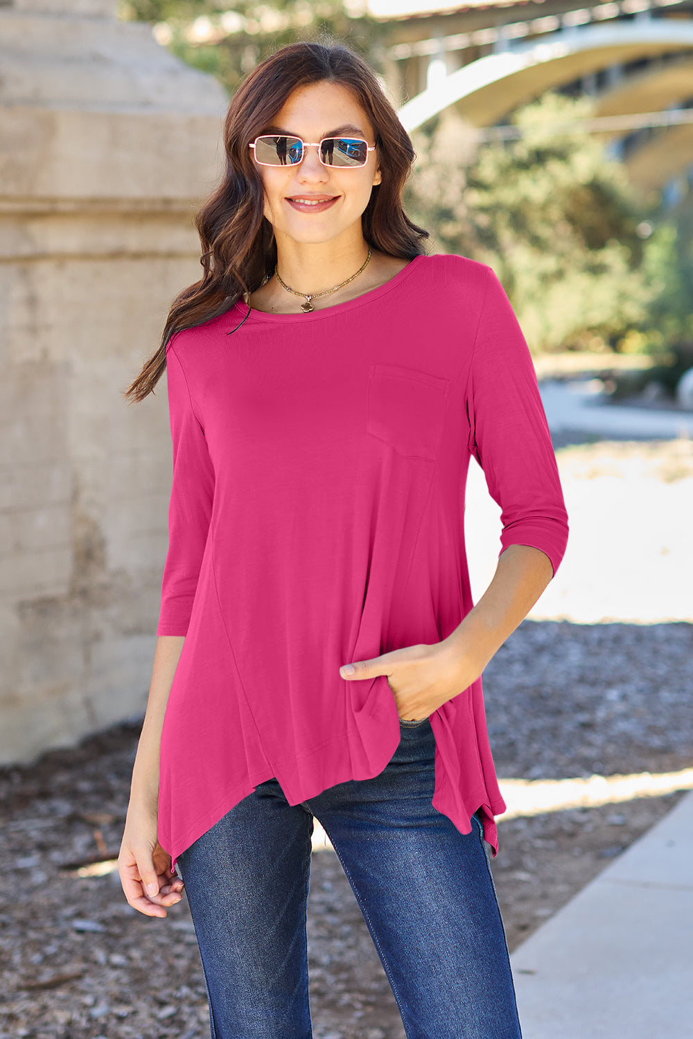 A woman stands outdoors wearing sunglasses, a Basic Bae Full Size Round Neck Pocketed T-Shirt with an asymmetrical hem and chest pocket made of rayon spandex, and blue jeans. She is smiling and has one hand in her pocket. Her basic style radiates effortless charm.