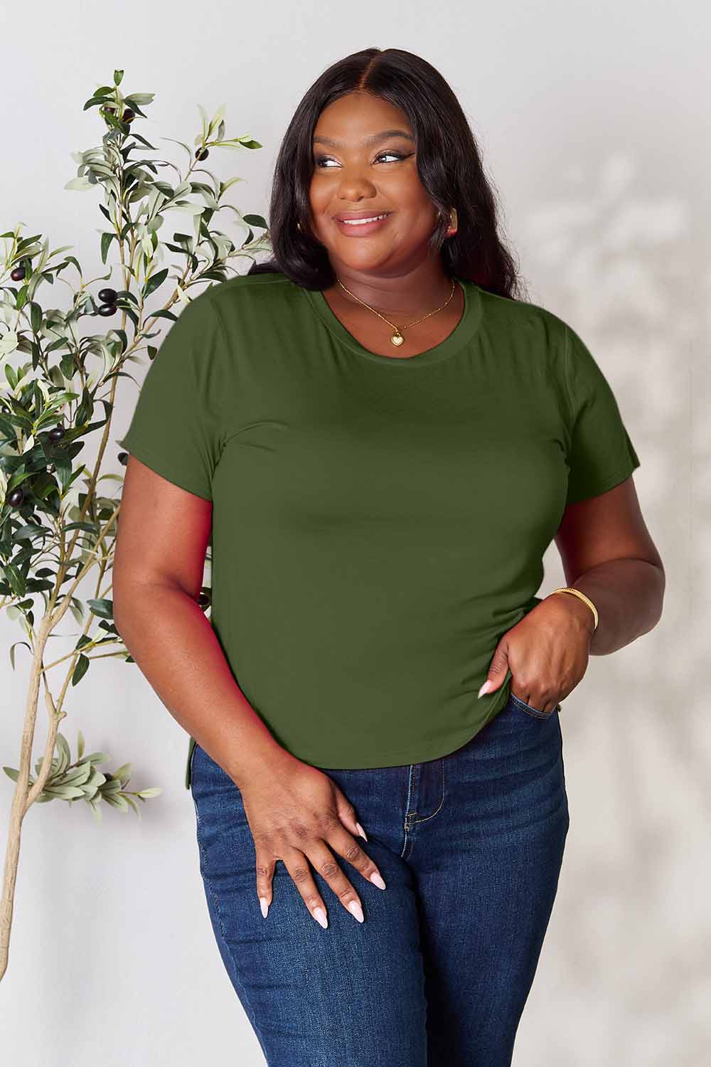 A woman stands next to an indoor plant, smiling, with one hand in her khaki pants pocket. In her pink Basic Bae Full Size Round Neck Short Sleeve T-Shirt and gold jewelry, she radiates casual elegance.