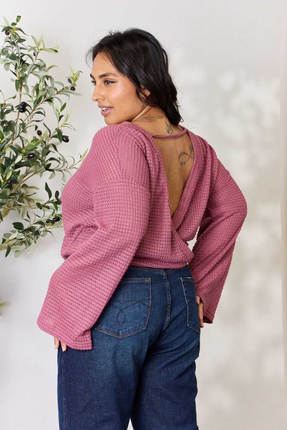 A person wearing a Culture Code Full Size Waffle-Knit Round Neck Long Sleeve Blouse in pink and jeans stands in front of a light background, complemented by a leafy plant on the left.