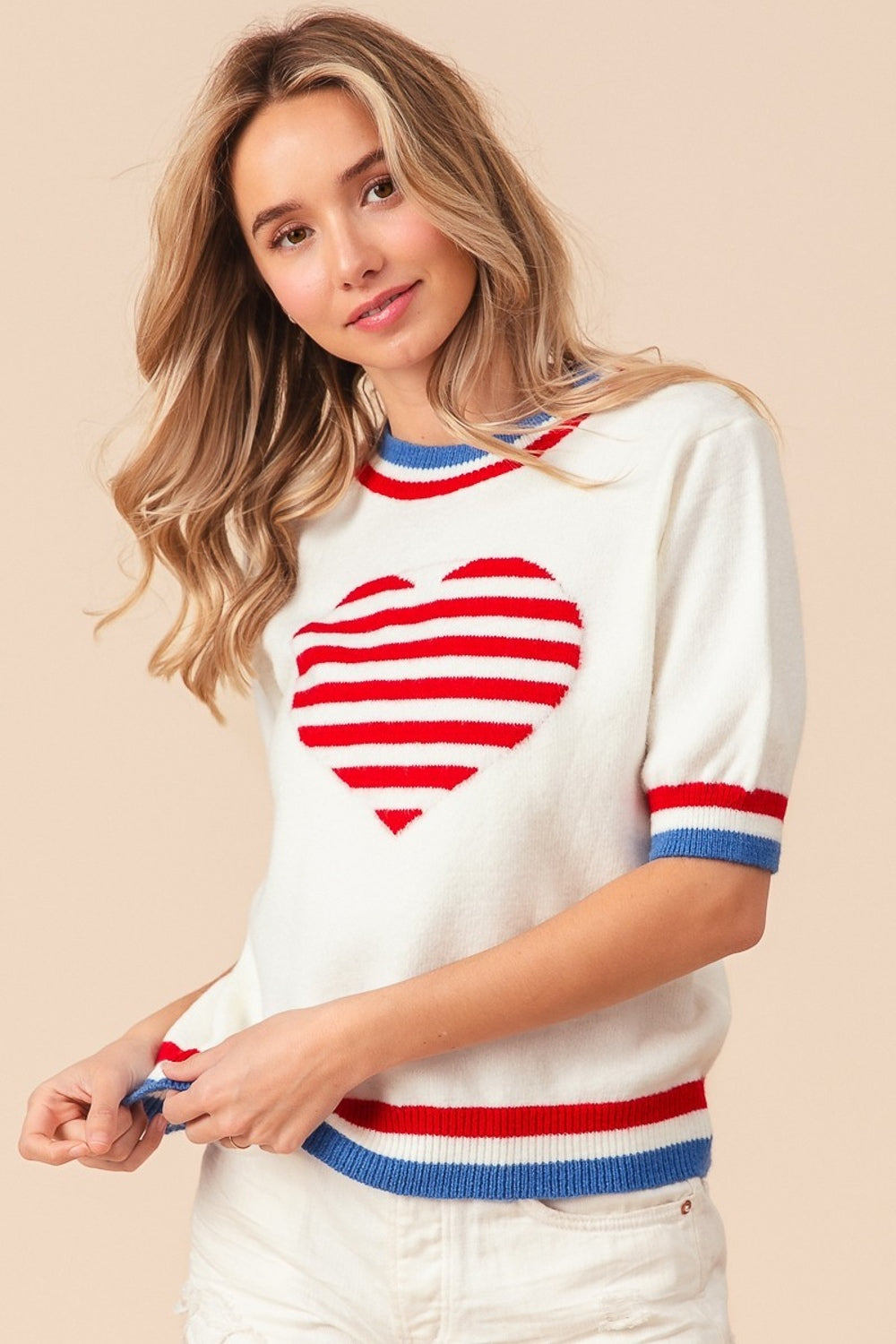 A person wearing the BiBi US Flag Theme Striped Heart Sweater, featuring a white short-sleeve design with red and blue stripes resembling the US flag and adorned with a red heart motif. They are also wearing white shorts and smiling with hands on hips.