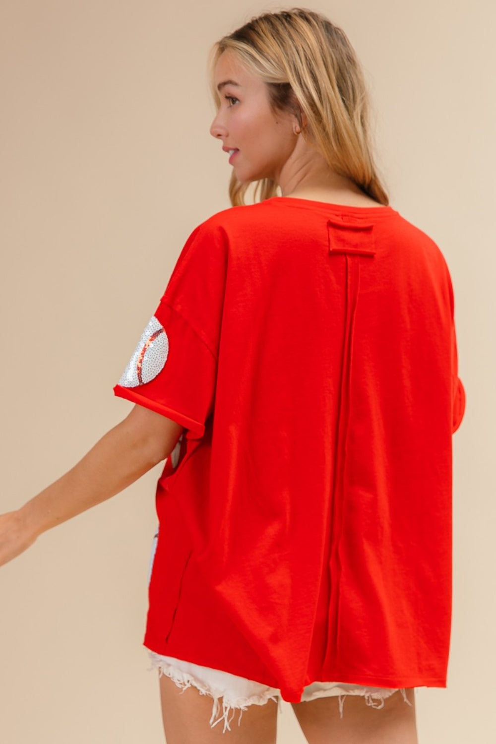 Woman wearing the BiBi Sequin Baseball Drop Shoulder Short Sleeve T-Shirt in red, paired with white shorts, posing against a beige background.