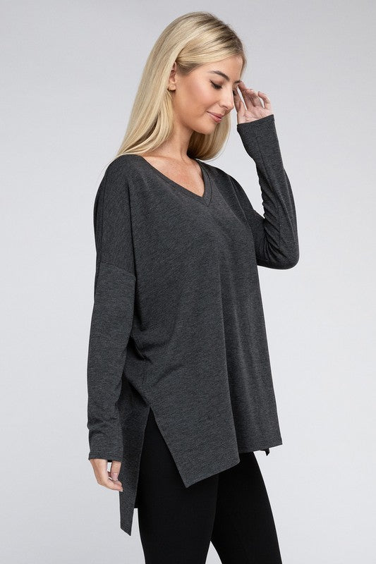 A woman with long, blonde hair is wearing a loose-fitting, brown Dolman Long Sleeve V-Neck Side Slit Hi-Low Hem Top and black pants. She poses with one hand on her hip and a neutral facial expression against a light grey background.