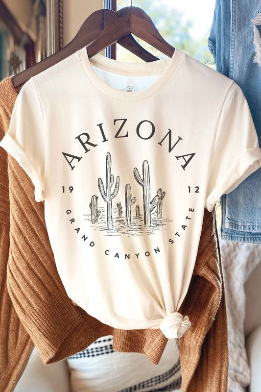 Person wearing an ARIZONA GRAND CANYON STATE Graphic Tee, made of premium 100% cotton with a design featuring the words "ARIZONA GRAND CANYON STATE 1912" and a drawing of cacti, paired with white shorts.