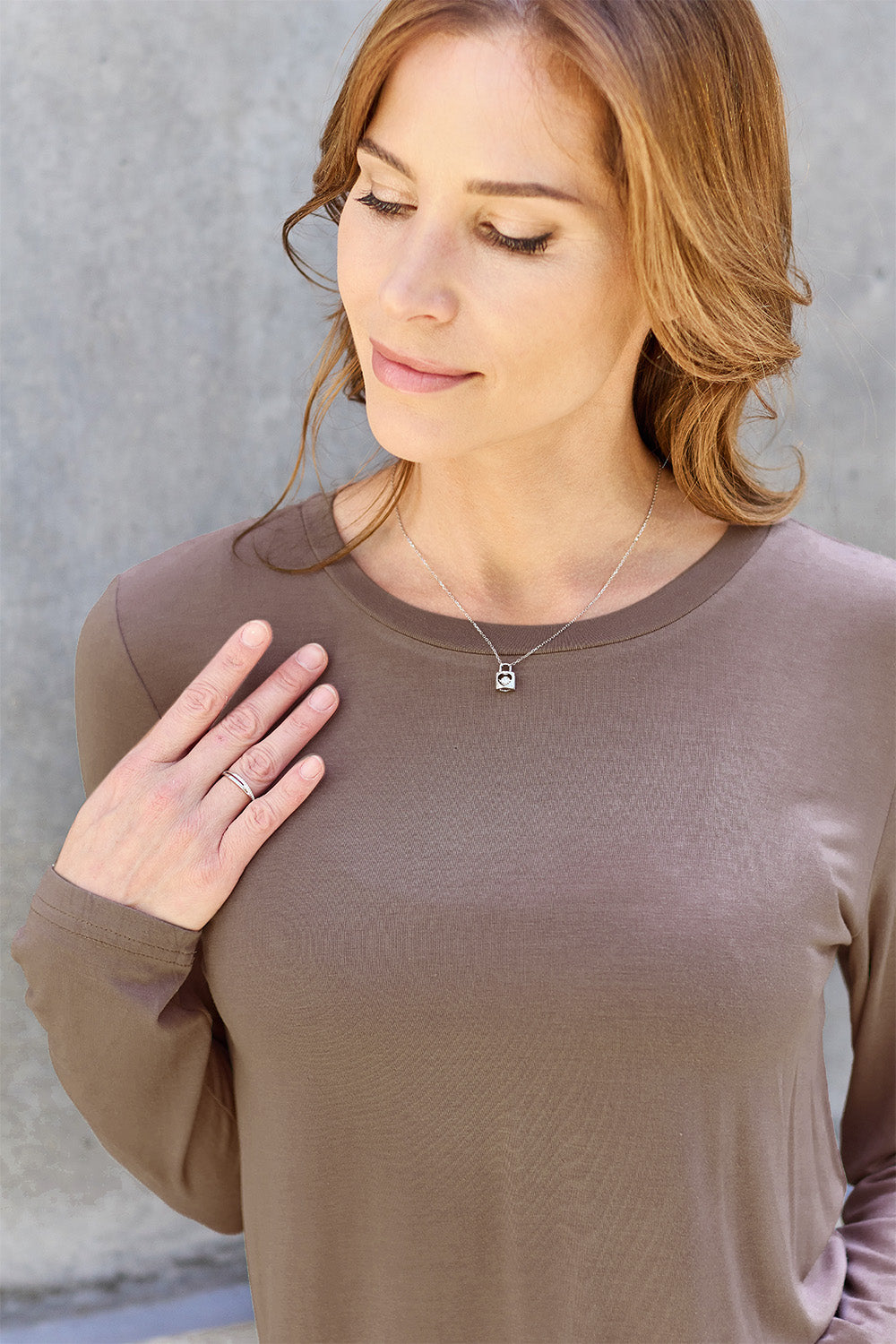 A woman with shoulder-length hair wearing the Basic Bae Full Size Round Neck Long Sleeve Top and blue jeans stands in front of a concrete wall, holding a beige handbag over her shoulder. Her outfit exemplifies basic style, with the top made from 100% polyester and recommended for machine wash cold.