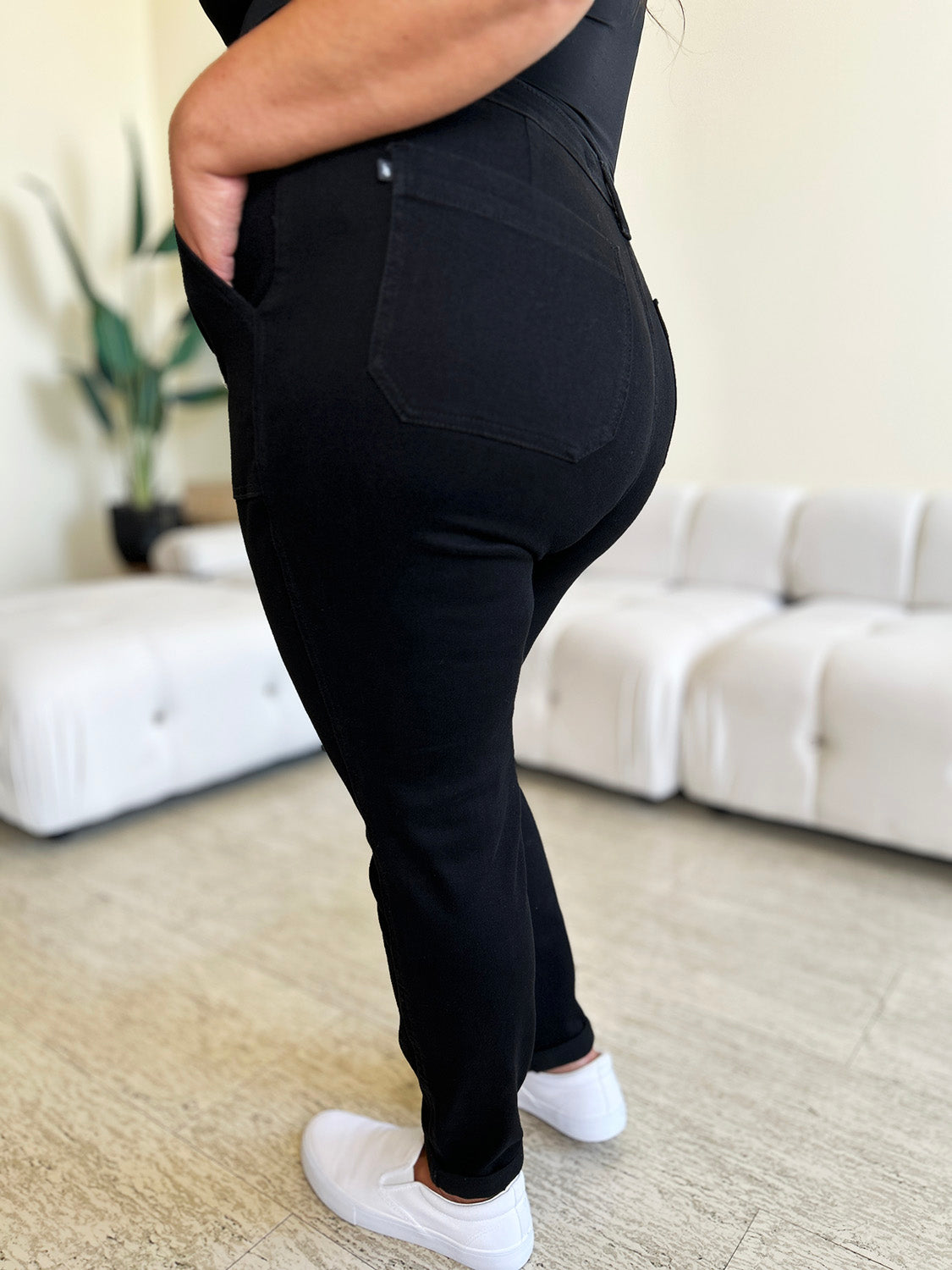 Person standing indoors, wearing a white top, black Judy Blue Full Size High Waist Double Roll Cuff Jeans, and white slip-on shoes, next to white couches and a plant in the background.