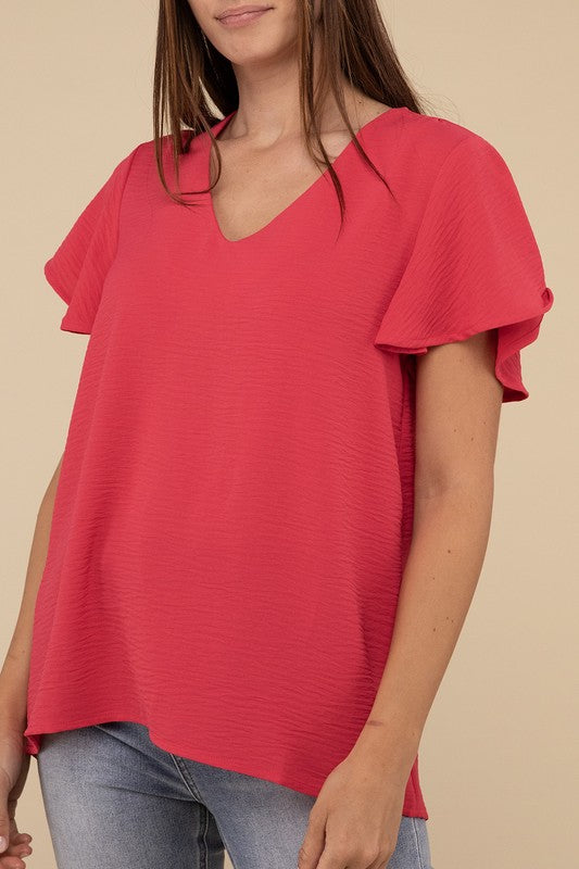 A woman with long straight hair models the Woven Airflow Flutter Sleeve Top in green, matched with light blue jeans, as she poses with her hands in her pockets against a plain beige background.