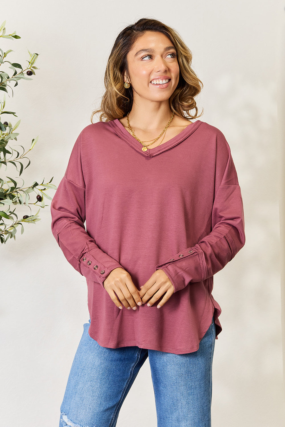 A person stands in front of a plain backdrop with a plant, wearing a maroon Culture Code Full Size V-Neck Exposed Seam Long Sleeve Blouse and blue jeans, smiling slightly with their hand on their hip. The relaxed fit of the polyester blend adds to the casual yet stylish look.