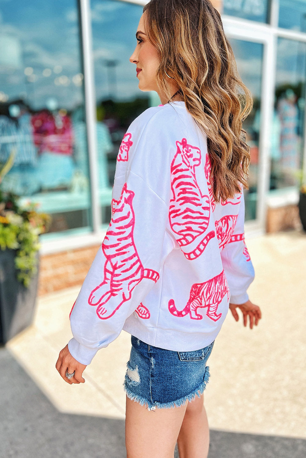 A person with long blonde hair wears a trendy Bright White Pink Animal Print Pullover Sweatshirt, paired with blue jeans, standing outdoors with their back facing the camera.