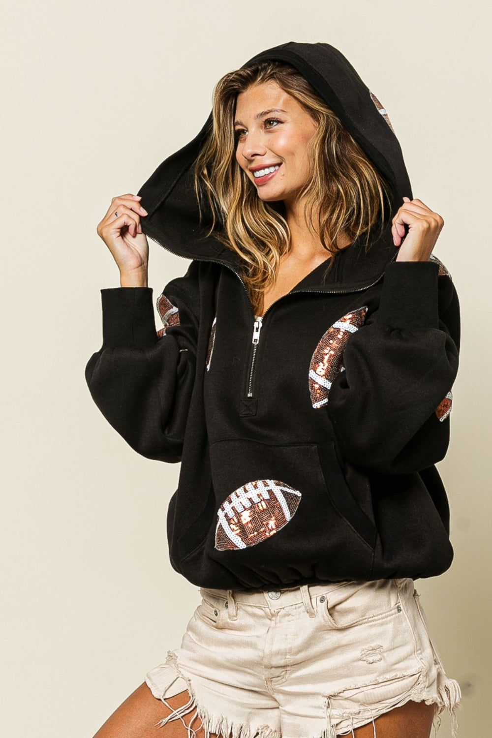 A person wearing a BiBi Sequin Football Half Zip Hoodie and beige shorts stands smiling in front of a neutral background with dried plants.